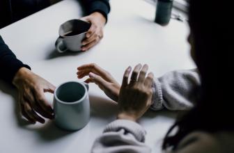 Tvær hendur sem mögulega tilheyra karlmanni hvíla á borði. Önnur þeirra heldur um kaffibolla og hin réttir annan bolla áfram. Tvær hendur sem mögulega tilheyra konu taka á móti bollanum.