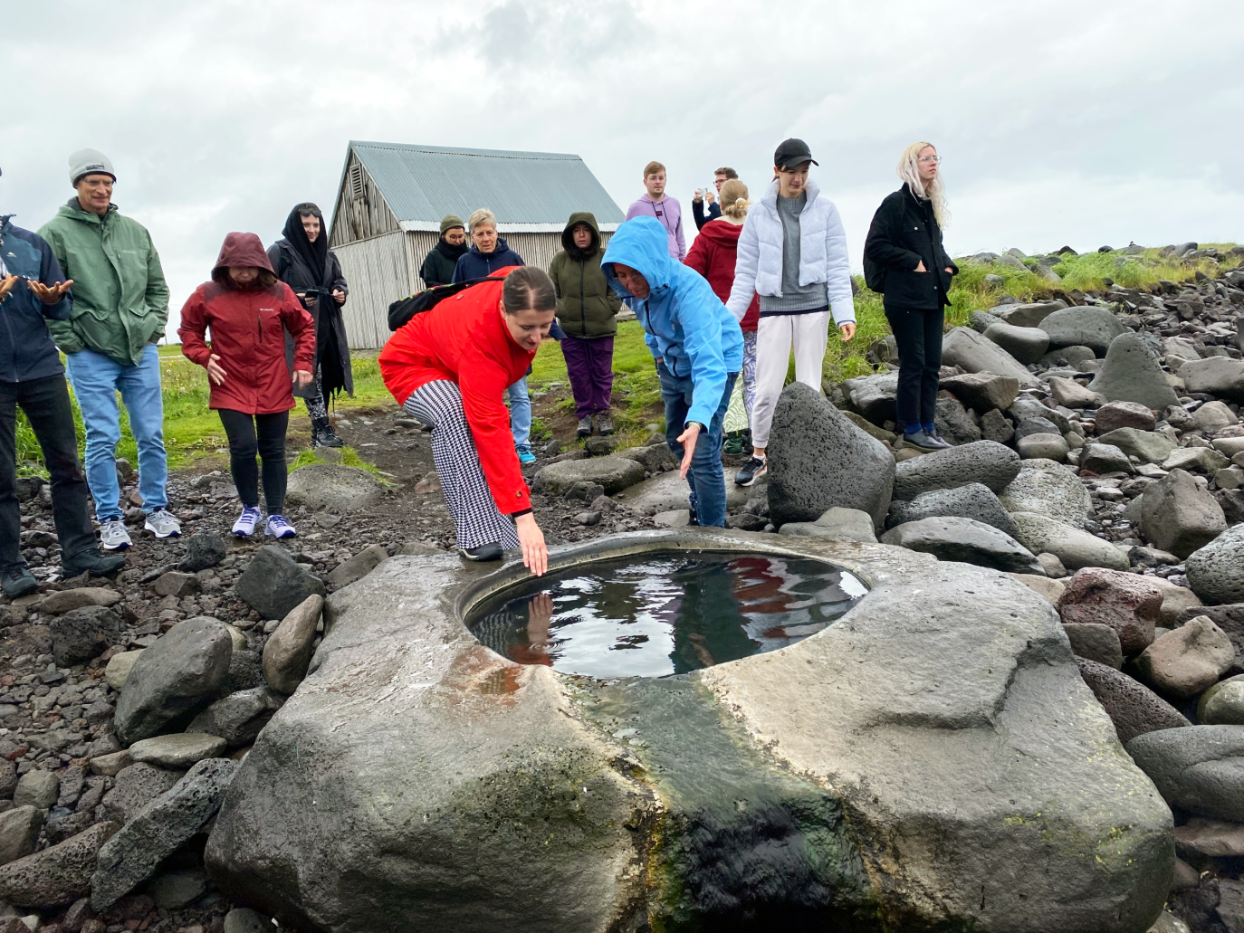 Hópur fólks virðir fyrir sér litla laug í stórum steini. Að baki er grár himinn.