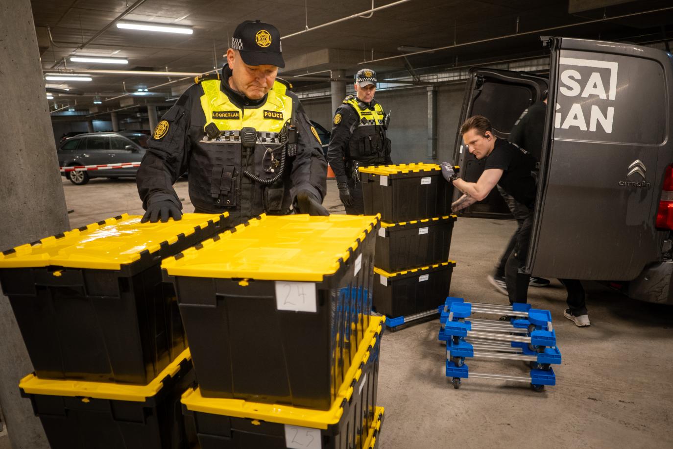 Grár, steyptur bílakjallari. Maður flytur stæðu af svörtum og gulum kössum úr svörtum sendibíl. Tvær stæður standa þegar á gólfi og tveir lögreglumenn standa álengdar.