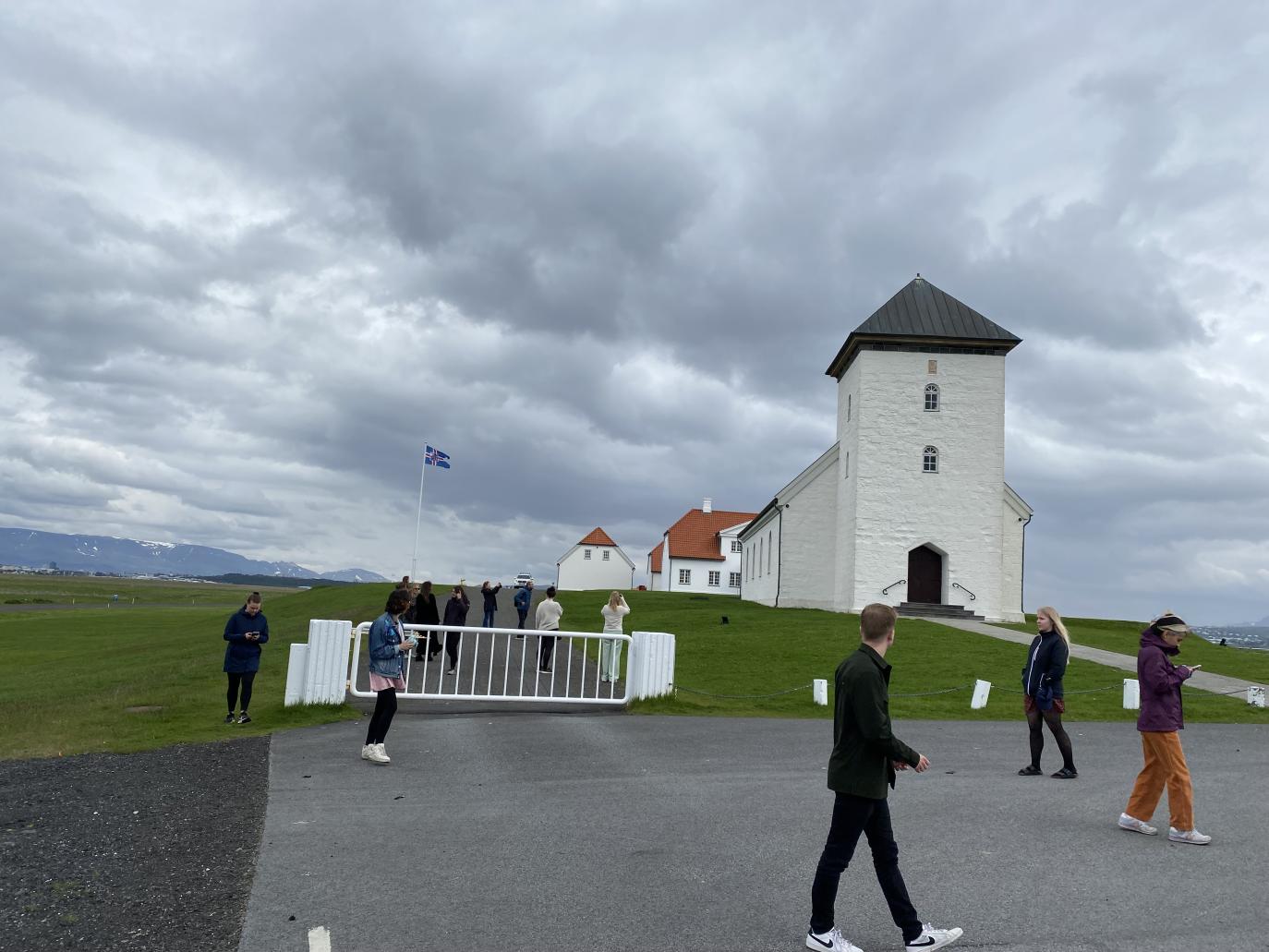 Hvítt, stórt, gamalt steinhús með breiðum turni. Fólk á vappi á bílastæði fyrir framan hvítt járnhlið. Fjær sér í stór, hvít, gömul hús með rauðu þaki og íslenska fánann dreginn að hún.