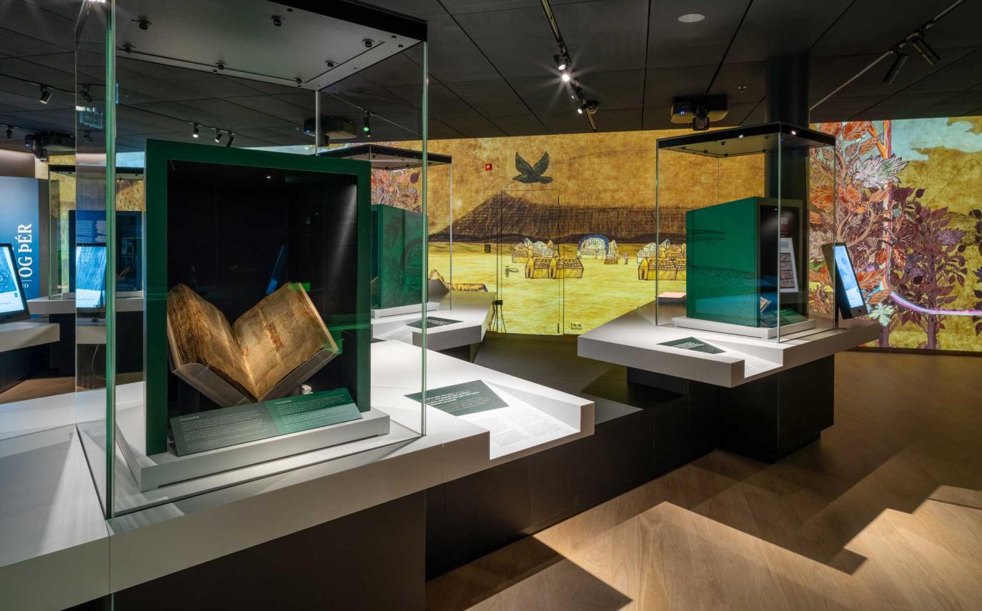 Overview inside exhibition space. On top of white tables are glass cases. Within them are green boxes that frame old manuscripts. The far wall is filled from floor to ceiling with drawn images of landscapes, foliage and a raven in flight.