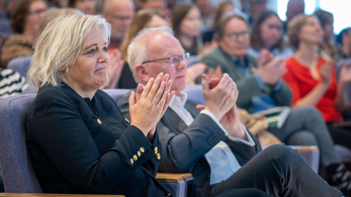 Fólk situr í sætum og klappar. Í forgrunni er kona í svörtum jakka og maður með gleraugu og í jakkafötum.