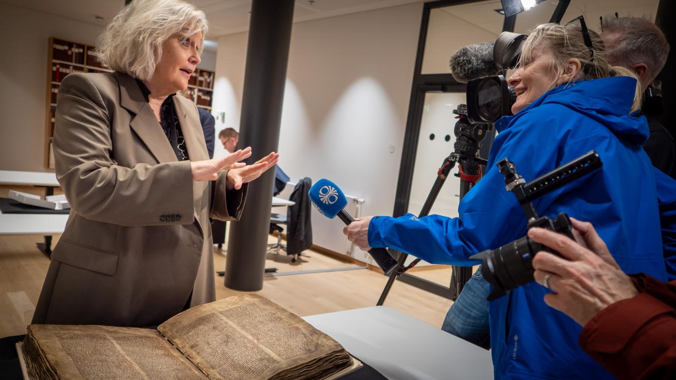 Kona stendur við handrit og talar við fjölmiðlakonu með hljóðnema. Einnig sést í myndavél.