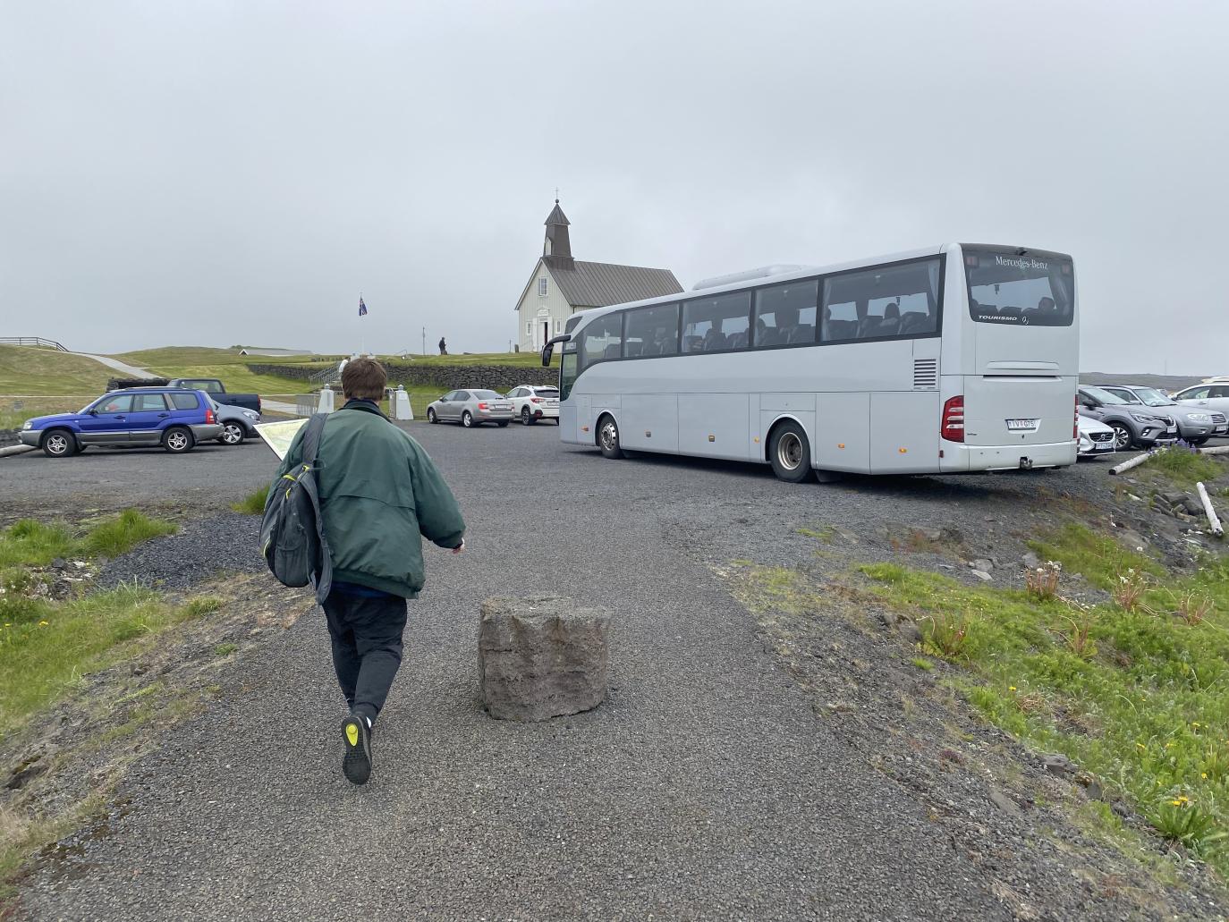 Manneskja gengur eftir göngustíg að bílastæði sem á eru margir bílar og stór rúta. Fjær sést gömul timburkirkja með mjóum turni.